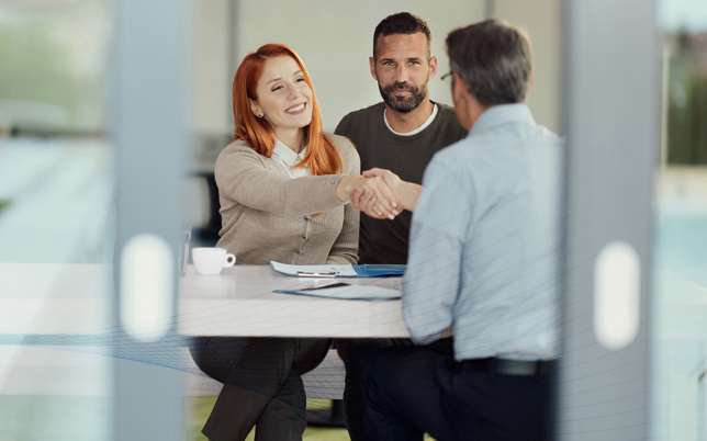 A couple sitting at a desk with F&I Manager, shaking hands, paperwork on desk.